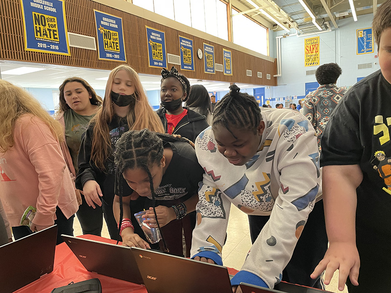 A group of five middle school students lean in to work on a few tablets.