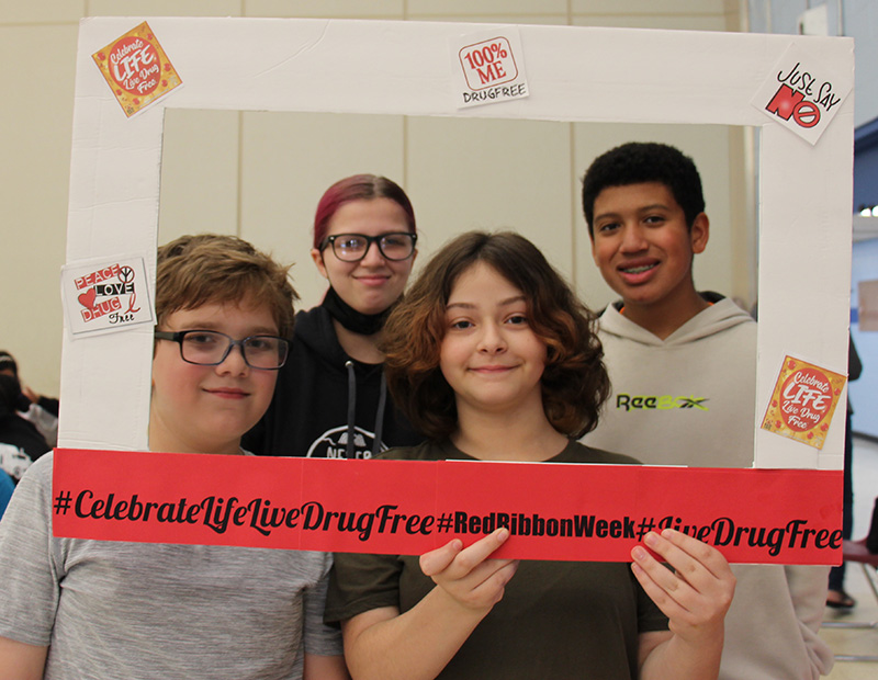 A group of four middle school students smile and hold up a large cardboard picture frame that says Celebrate life Live Drug Free.