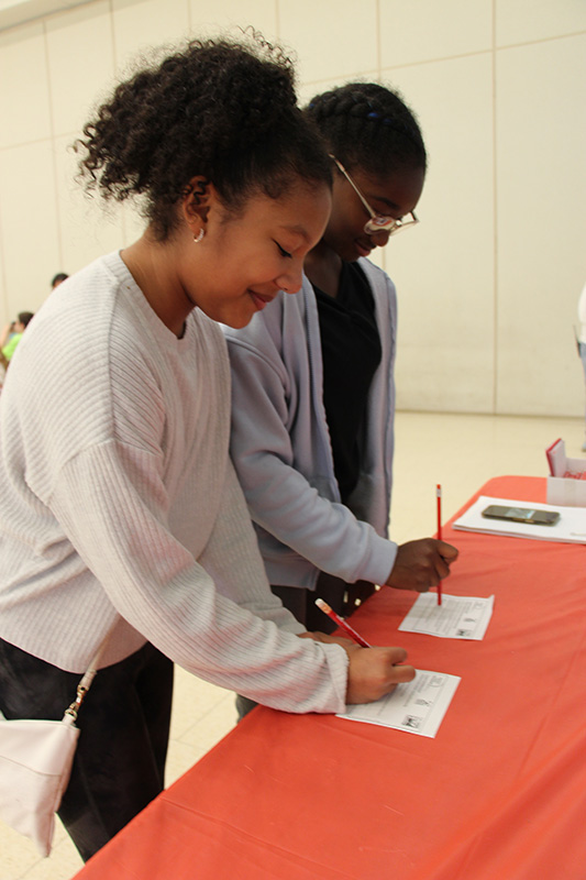 Two middle school girls sign pieces of paper.