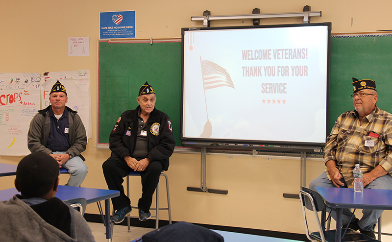 Three men, each wearing a cap indicating they are veterans, sit on chairs in the front of a classroom. There is a screen by them with an American flag that says Welcome Veterans! Thank you for your service.