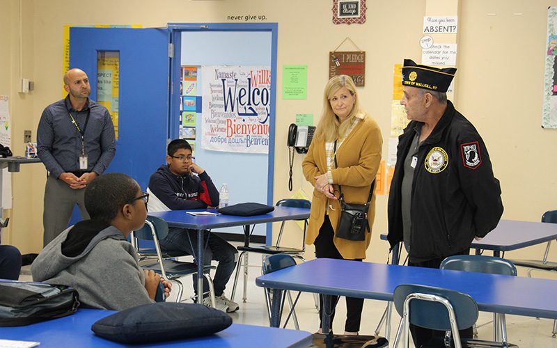 An older man wearing a blue jacket and blue cap stands in a classroom talking to a middle school student who is sitting at a desk. Two adults stand and listen and another students sits at another desk listening.