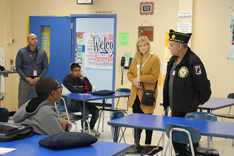 An older man wearing a blue jacket and blue cap stands in a classroom talking to a middle school student who is sitting at a desk. Two adults stand and listen and another students sits at another desk listening.