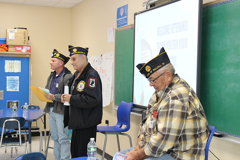 Three older men, all wearing veterans caps, stand and sit in front of a classroom of students. There is a screen in between them.