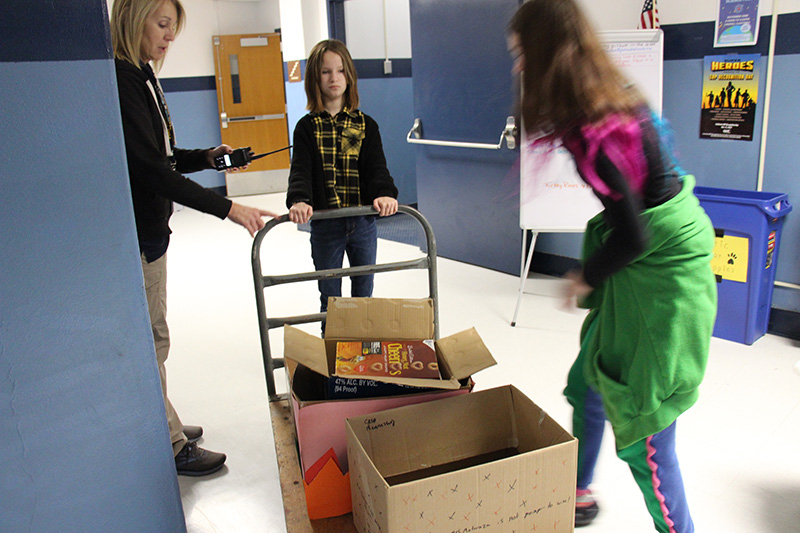 Two middle school students and an adult wheel a dolly with boxes of food on it.