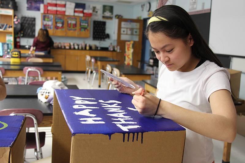 A middle school student, with long dark hair held back with a headband, paints Happy Thanksgiving on a blue box.