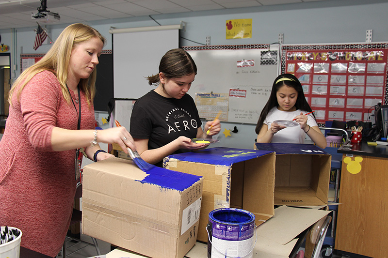 Two middle school girls , both with dark hair, and a woman with long blonde hair, paint boxes that say Happy Thanksgiving.