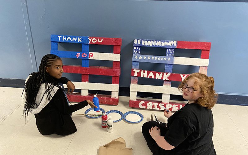 Two middle school girls sit on a floor and turn around to look at the camera. They are painting pallets to look like American flags.