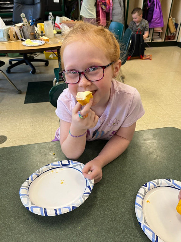 A girl with red hair pulled back in a ponytail, wearing a light colored shirt and glasses holds a piece of cornbread with butter on it as she takes a bite.