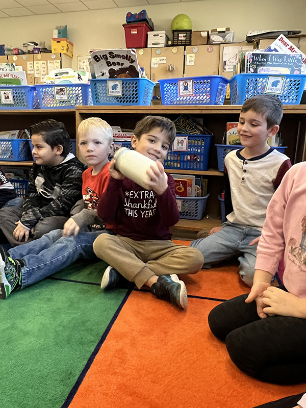 A group of four elementary students sit on a multi-colored rug. The boy in the center is shaking a jar filled with cream.