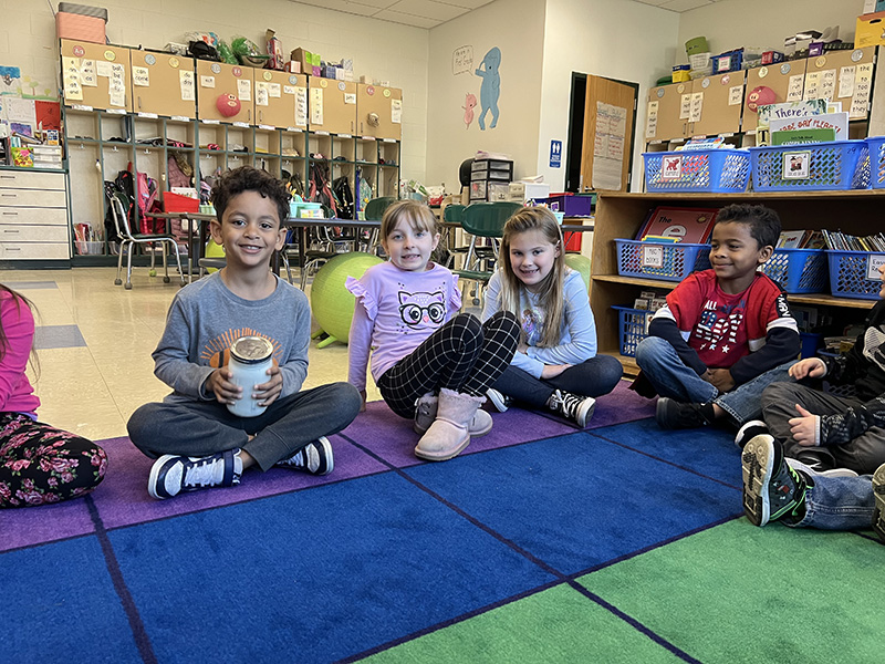 A group of four students sit on a multi-colored rug. The boy on the left is holding a jar filled with creamy butter.