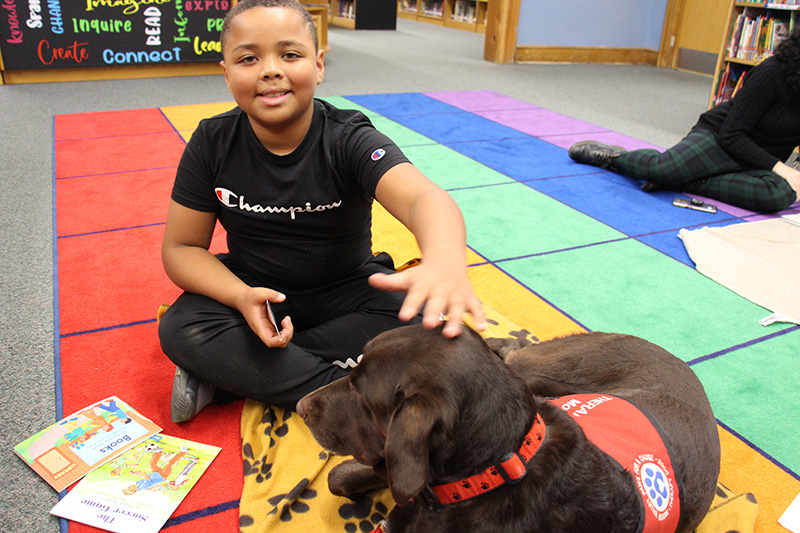A boy with short dark hair smiles as he sits on a colorful rug. He is petting a large chocolate lab.
