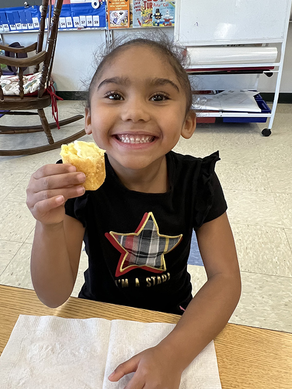 A little girl whose dark hair is pulled back into a ponytail smiles broadly as she holds up a piece of cornbread with homemade butter.