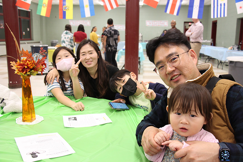 A family of five sit at a round table with a green tablecloth on it. The elementary age girl on the right has long dark hair and is wearing a black and white striped shirt ad a white mask. The woman next to her is smiling. There is an elementary age boy next to her giving a peace sign and wearing a black mask. A man is next to him, wearing glasses and a dark shirt. He is smiling and holding a young child.