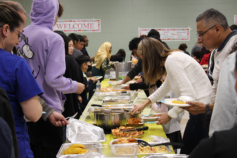 A long table with many containers of food on it. There are people lining each side of the table getting food.