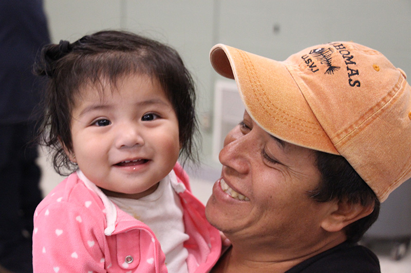 A man wearing a peach-colored baseball cap holds a little girl about 2 years old with dark hair. She is wearing a pink jacket and is smiling.