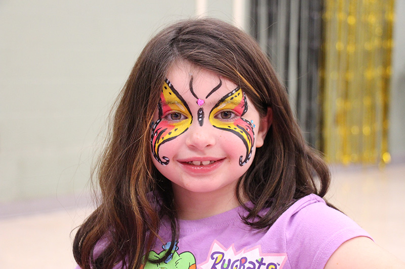 An elementary-age girl with long dark hair, wearing a light purple shirt, smiles. She has a large butterfly painted in orange and yellow and black on her face.