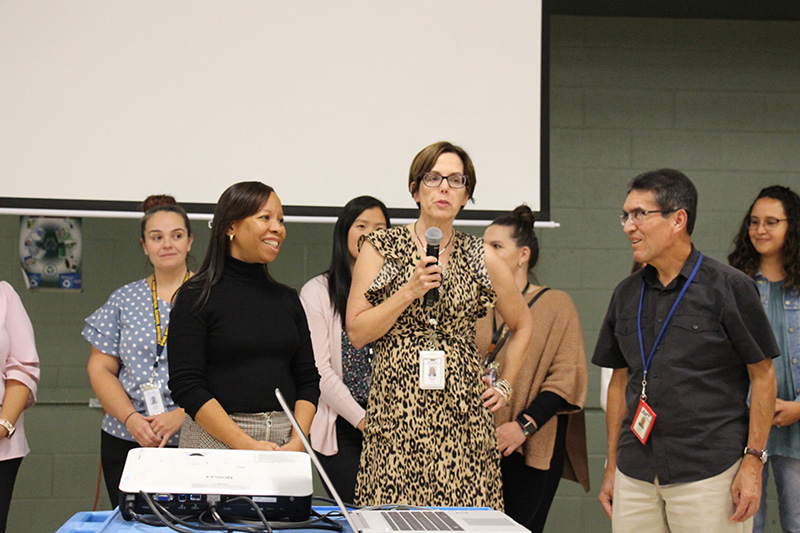 A group of women and one man stand together. One woman is holding a microphone. A woman next to her is smiling.