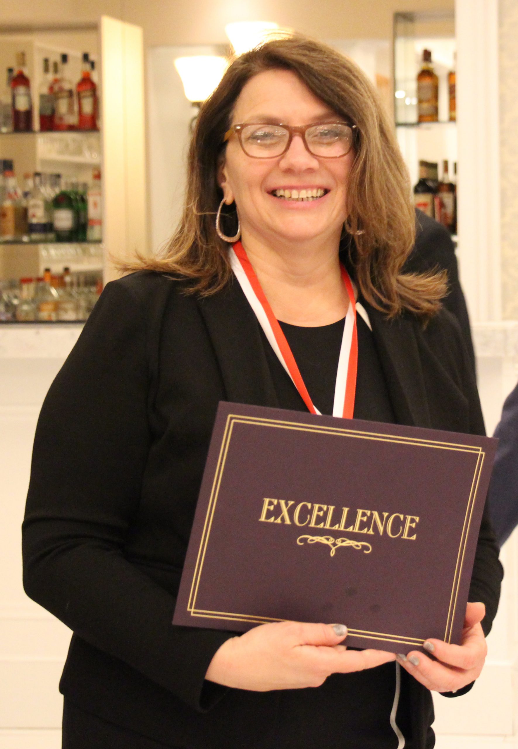 A woman with longer dark hair smiles. She is wearing a black dress, glasses and is holding a certificate that says Excellence on it. She also has a medal around her neck on a red, white and blue ribbon.