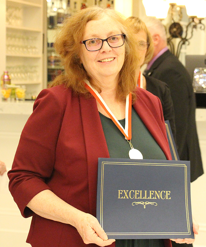 A woman with shoulder-length reddish hair holds a certificate in front of her that says Excellence. She is wearing a black shirt, maroon sweater and has a medal around her neck.