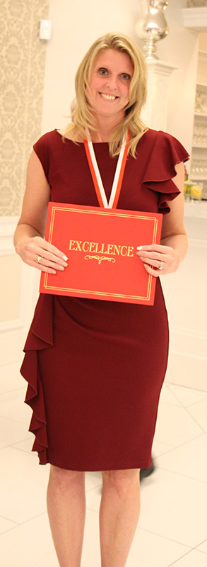 A woman with shoulder-length blonde hair, wearing a maroon dress, smiles and holds a red certificate in front of her that says Excellence. She has a medal around her neck.