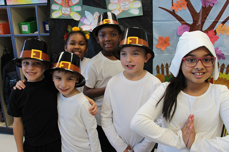 A group of six second-graders, four with pilgrim hats, one with a ring of yellow flowers and one with a white pilgrim bonnet. They are all smiling.