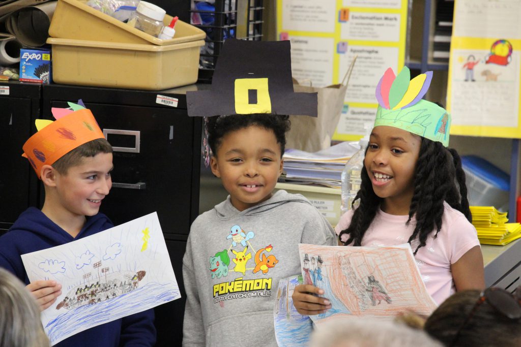 Three second-grade students, dressed with paper hats - two with feathers and one a pilgrim hate - stand together smiling. The girl on the right is speaking.