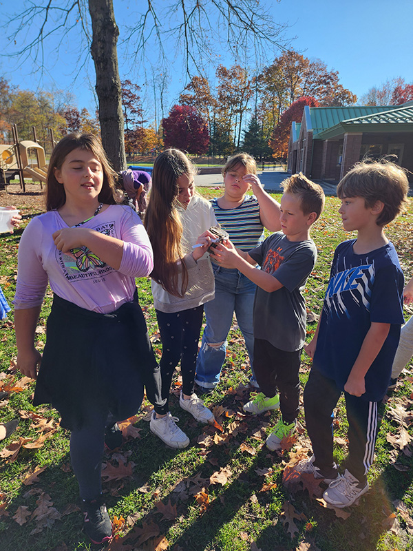 Five older elementary students are outside collecting things from nature. There is blue sky and lots of leaves on the ground.