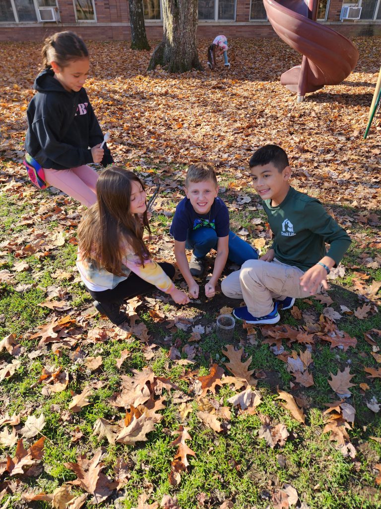 Four elementary students bend to the ground to pick up natural matter for an experiment. The ground is covered in leaves.