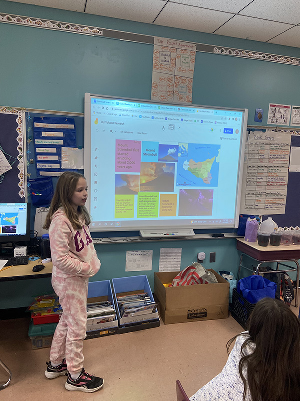 A student stands in front of a class in front of a white board where there is a presentation displayed.