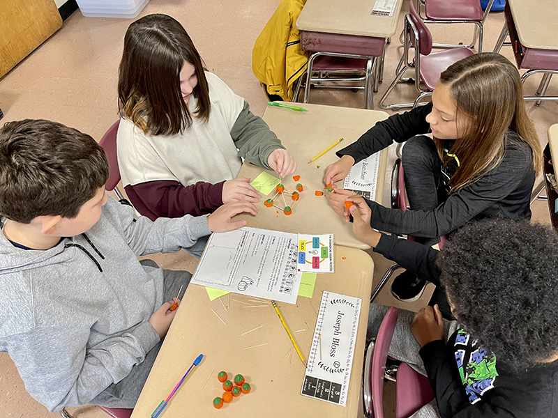 A group of four fifth grade students sit at a table and work on a project with toothpicks and pumpkins. The view is above the table as they work.
