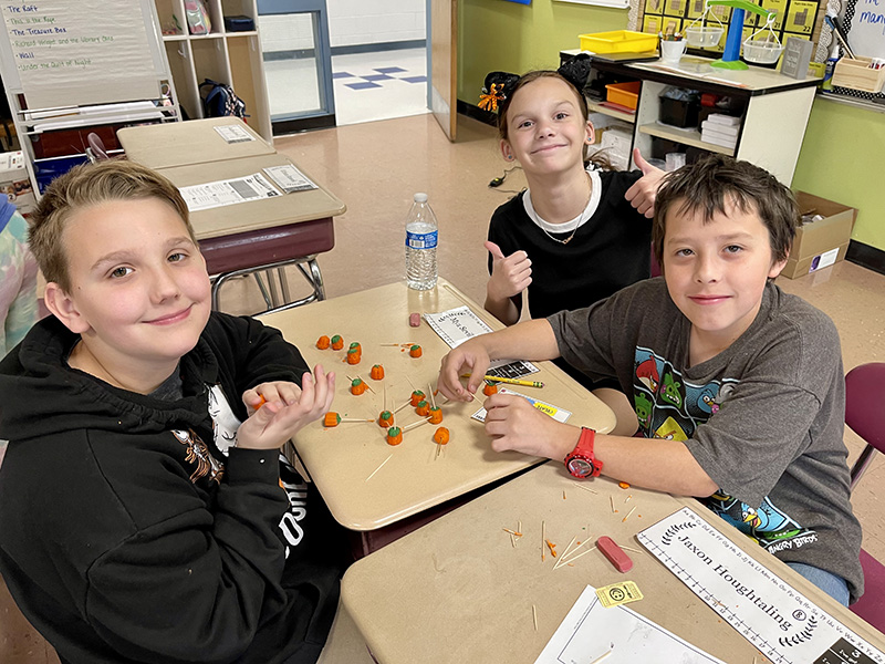A group of three fifth grade students sit at a table and work on a project with toothpicks and pumpkins. One student is giving a thumbs up.