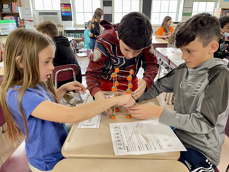 A group of three fifth grade students sit at a table and work on a project with toothpicks and pumpkins.