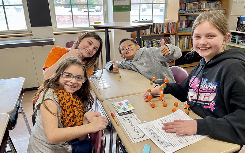 A group of four fifth grade students sit at a table and work on a project with toothpicks and pumpkins. One student is giving a thumbs up. they are all smiling.