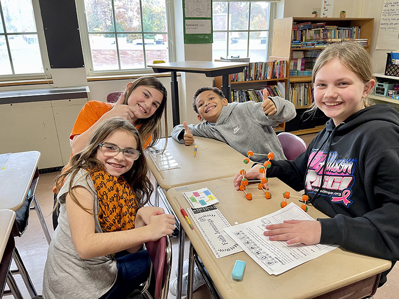A group of four fifth grade students sit at a table and work on a project with toothpicks and pumpkins. One student is giving a thumbs up. they are all smiling.