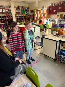 Two elementary students stand at a red line on the floor and toss pencils.