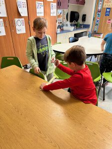 Two elementary age boys  are at a table holding a ruler on the edge of it.