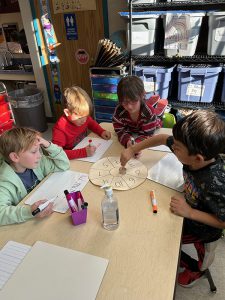 Four elementary students sit around a table with a round disk in the center with numbers on it. They are spinning a top on it.