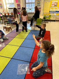 Two elementary students stand on a multi-colored rug with sticks looking like they are fishing.