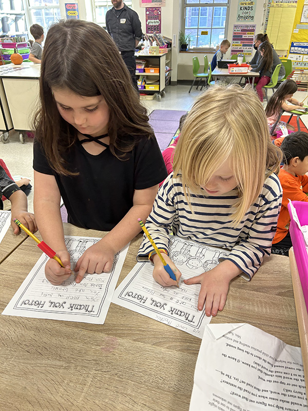 Two first-grade students work on papers thanking  veterans. The girl on the left has long dark hair and is wearing a black shirt. The child on the right has long blonde hair and is wearing a black and white striped shirt.