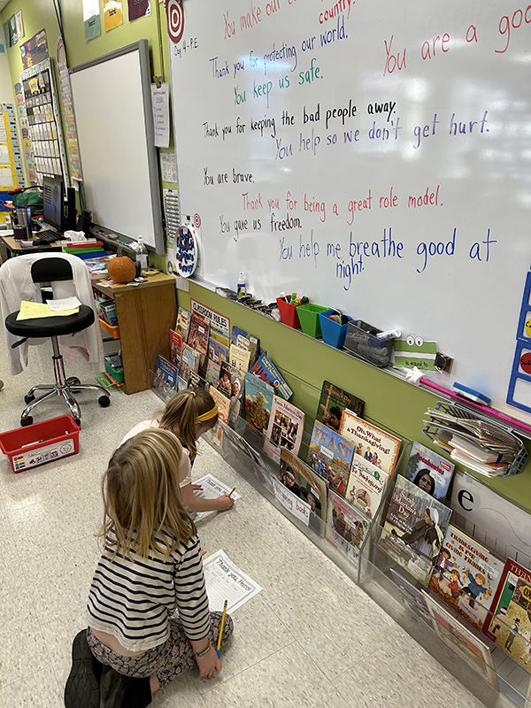 Two first-grade students sit on the floor looking up at a white board, as well as a rack of books underneath it.