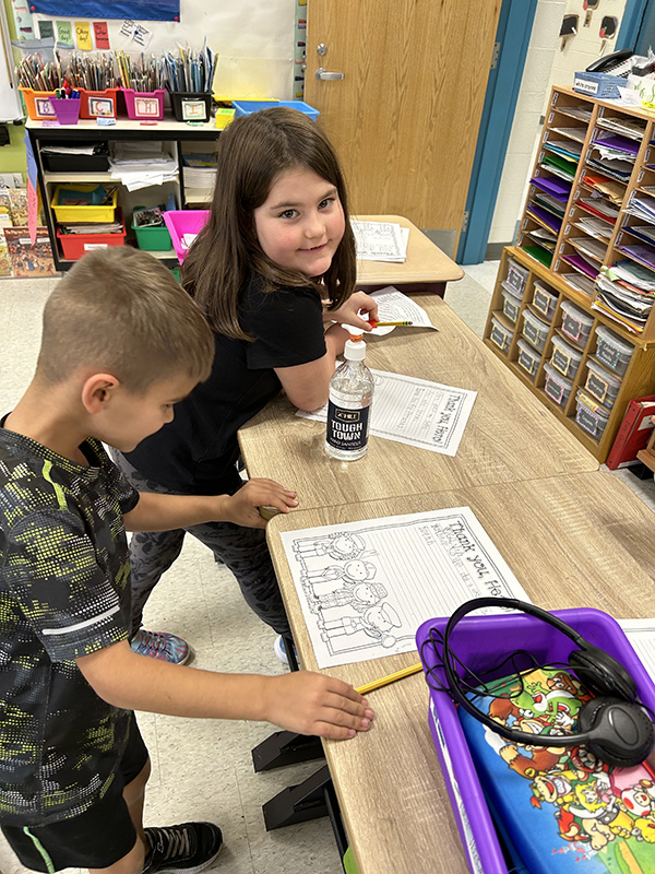 Two first-grade students work at a table. The student at the top has a black shirt on and long dark hair. The boy next to her has short hair and is looking at the paper on the table.