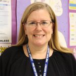 A woman with long blonde hair and glasses smiles. She is wearing a black shirt with a blue lanyard around her neck that says Pine bush Central Schools.