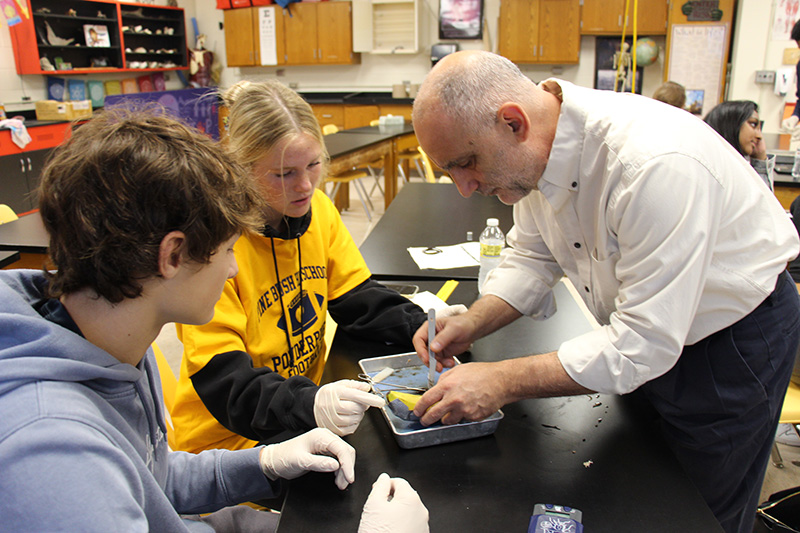 A man in a white shirt and dark pants stands on the right, bending over and looking into a tray with two high school students who are on the right. He has forceps in his hand as he moves something.