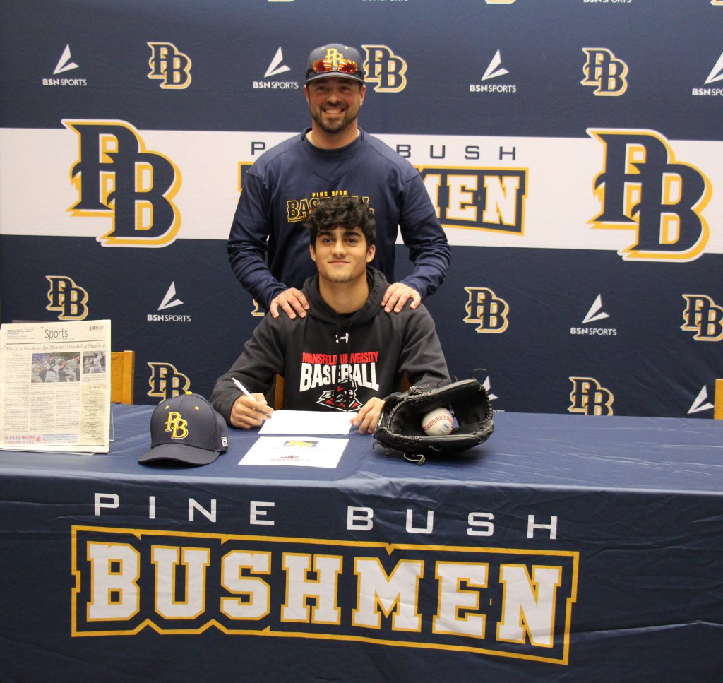 A high school young man sits at a table with a pen in his hand and paper in front of him. There is a baseball cap and a baseball mitt and ball on the table. Behind him is a man with his hands on the young man's shoulders. The tablecloth says Pine Bush Bushmen. The backdrop does too.