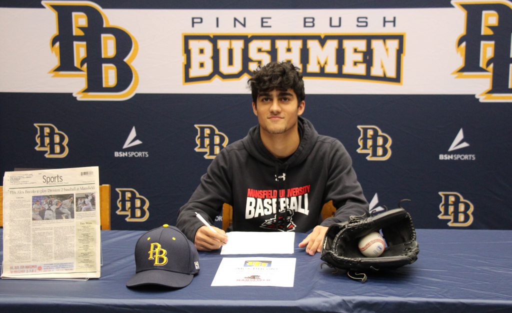 A young man with short dark hair sits at a table and signs a piece of paper. On the table is a baseball cap that says PB and a mitt and ball. He is wearing a shirt that says Mansfield baseball.