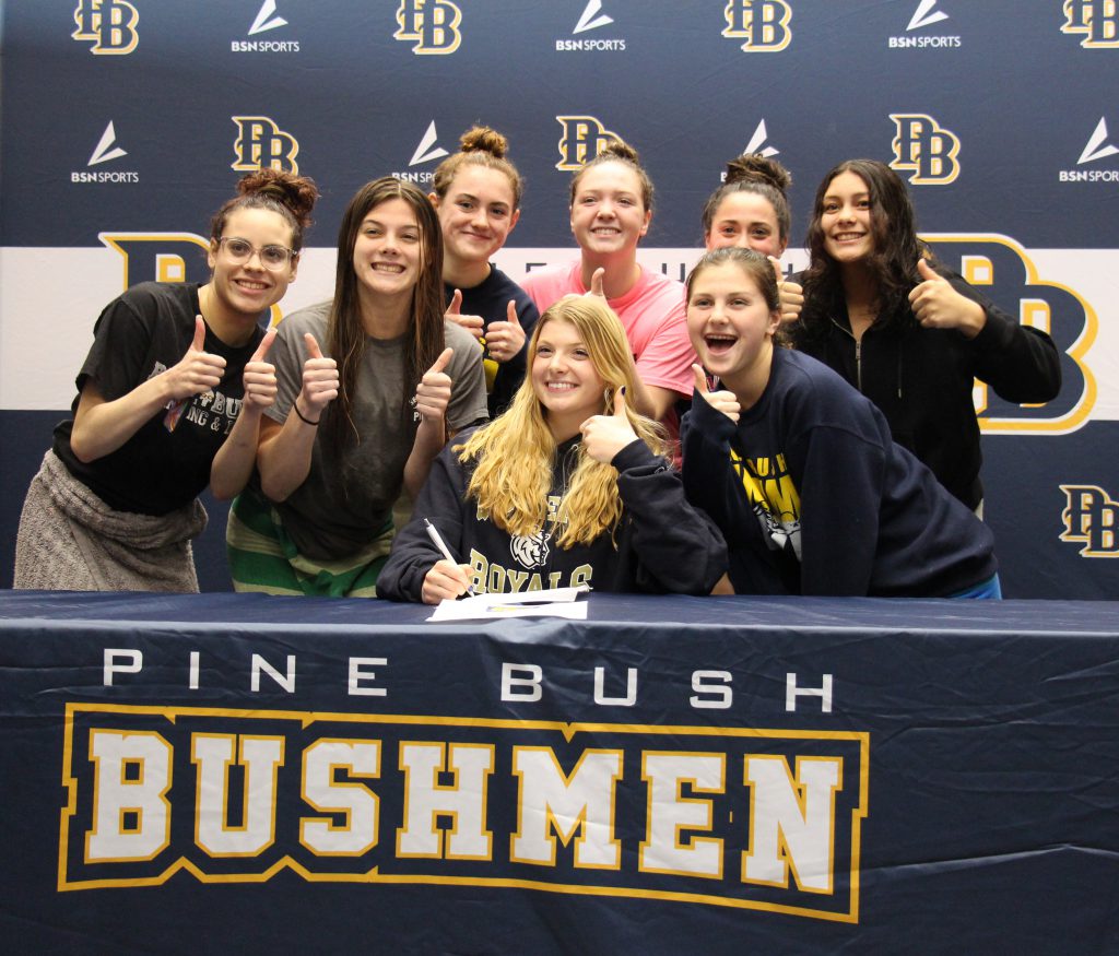 A large group of high school girls stand around another high school girl with long blonde hair who is signing a piece of paper. They are all giving a thumbs up. The table cloth sayd Pine bush Bushmen.