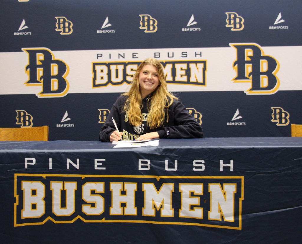 A high school age young woman with long blonde hair sits at a table and signs a paper. She is smiling broadly. Behind her is a backdrop that says Pine Bush Bushmen PB.