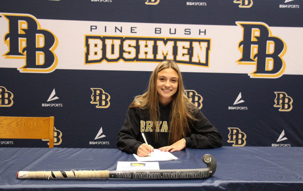 A young woman with long blonde hair sits at a table and signs a piece of paper. Behind her is a large backdrop that says Pine Bush Bushmen PB. There is a field hockey stick on the table.