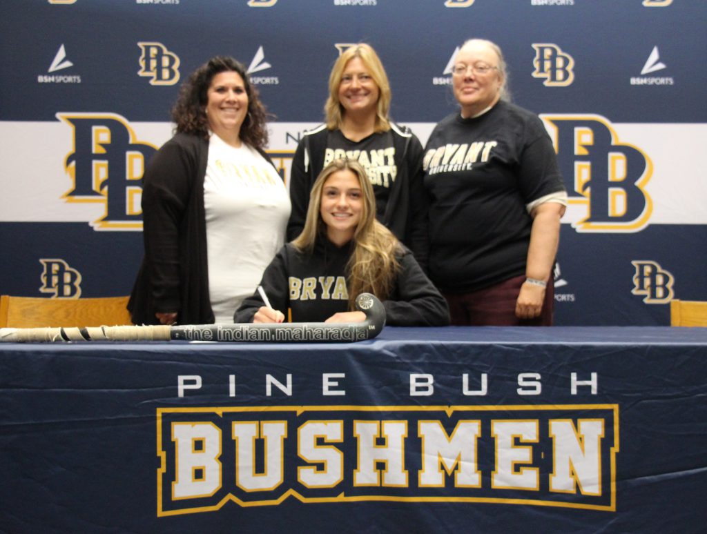 A young woman with long blonde hair wearing a navy blue sweatshirt that says Bryant on it, sits at a table and smiles. Behind her are three women smiling. The tablecloth says Pine bush Bushmen.