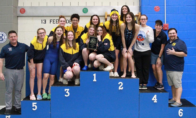 A group of about 13 high school girls in blue and gold warm-ups and four adults stand on a swimming platform numbered from 1 to 7.